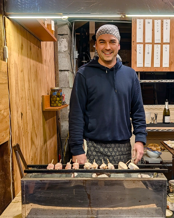 A man smiling standing behind a grill with skewers on it